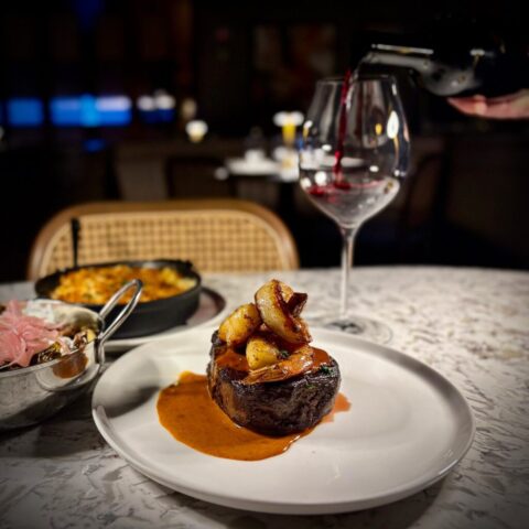 A delicious steak is set on a white plate, it is topped with scallops and shrimp dripping in a rich creamy red sauce. In the background a glass of red wine is slowly being poured.