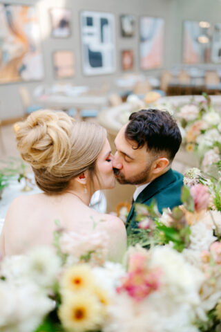 A wedding shot of a couple kissing at the Dean Restaurant. The man is in a dark greens suit and he has dark brown hair and a beard. The woman is blond with her hair styled up into a pretty bun and is in a white gown. The couple is surrounded by blurry white and pink flowers, the large glamorous art of the dean can be seen in the background.