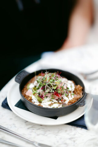 A black skillet filled with brisket and topped with white cream and micro greens is centered on a white plate.