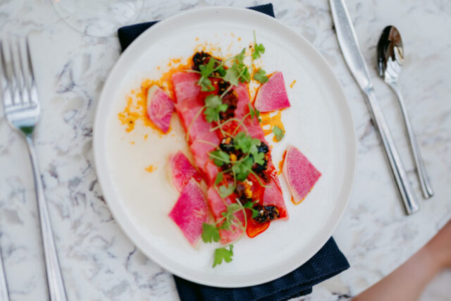 A bright pink beet salad is topped with cilantro and centered on a white plate. The plate sits on an elegant white and black marble table with a silverware place setting around it.