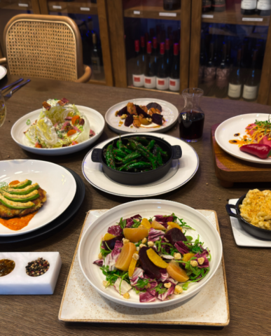 Appetizers are spread out on a wooden table. each appetizer is carefully placed on a white plate for a bright and clean presentation of dishes. There are a variety of sides including multiple fresh salads, macaroni, and crab cake.