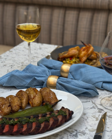 A white marble table is set for two. On the table two pretty blue napkins with golden rings sit in the middle separating a plate of steak and chicken. Two glasses of wine one red and the other white are on the edges of the table.