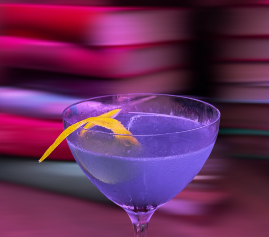 A funky purple cocktail topped with a bright yellow lemon peel is centered in this photo. Behind the glass a swirling riot of colorful books is the background, creating a feeling of chaos, fun, and excitement.
