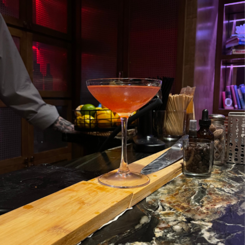 A red cocktail in a wide cocktail glass sits perched on a block of wood. The bar beside it is swirling with browns and blacks. The background showcases the variety of custom cocktail ingredients used allowing the viewer to see the level of care that goes into each drink.