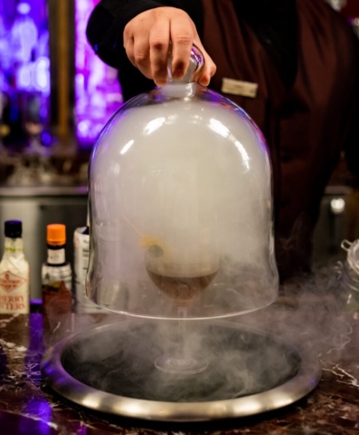 A large glass cloche is being lifted off of a tray, a heavy mist is seeping out of the glass enclosure and spreading around the bar top giving a mystical and beautiful atmosphere. As the smoke dissipates a custom craft cocktail is revealed to be on the tray.