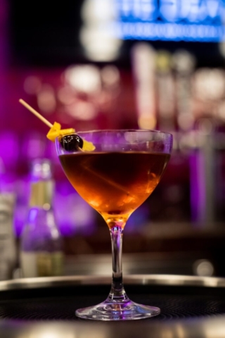 A cocktail is centered in the image. It's colors is a dark brown that turns into a glowing amber at the bottom. It is topped with a orange slice and blackberry. The background is a colorful and blurry display of lights giving off the image of a lively atmosphere where one can order fun fruity drinks.