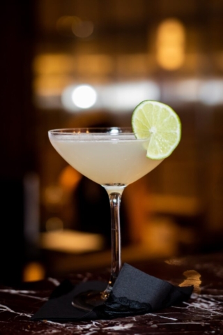 A thin glass cocktail is served on a black napkin. The color of the drink is a soft golden taupe and the drink features an accent of lime on the rim giving it a chic appearance