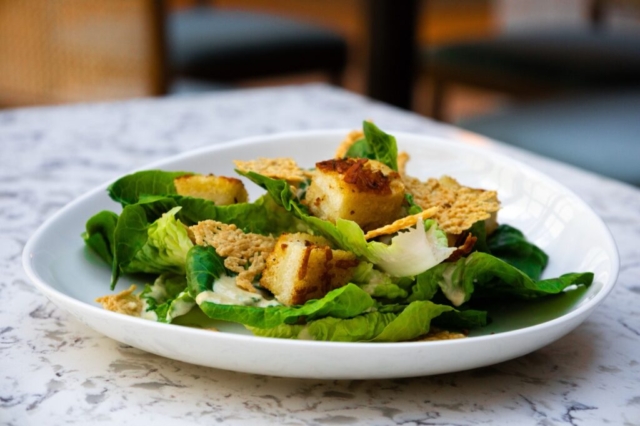 A close up shot of a cesar salad. The baby gems leaves are crisp and green. The salad is topped with freshly made parmesan crisps and Texas toast croutons.