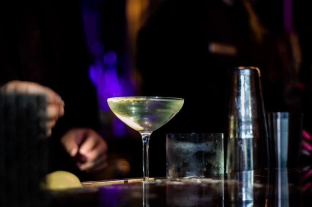 A futuristic style shot of a golden clear cocktail swirling in a glass. The condensation of the cool drink is visible to the eye, and the background is blurred and moody with lights reflecting off of silver drink shakers. The ambience is cool and intriguing.