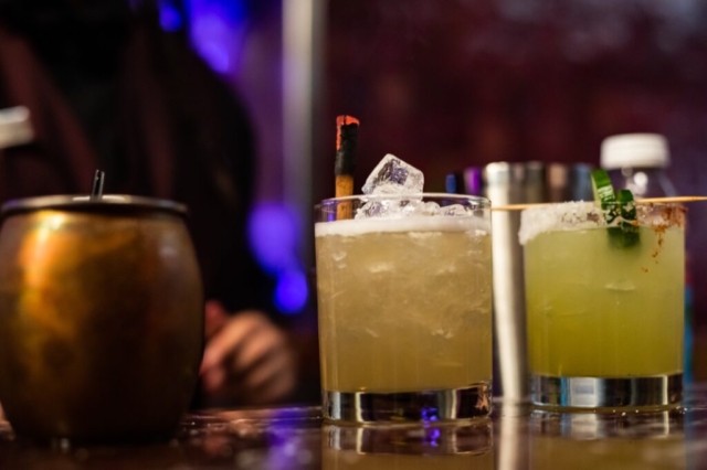 A close-up photo of three drinks on a bar counter. Two short cocktail glasses filled with ice and garnishes ( one margarita with cucumbers and jalapeno, and the other garnished with cinnamon stick) sit in the foreground, while a copper mug is visible to the left. The background is dimly lit with hints of blue light, providing a upscale atmosphere at the Dean.