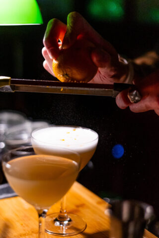 A moody shot of a bartenders hand zesting an orange onto a foamy drink below. The drinks are standing on a wooden cutting board and lit up by a vintage bankers lamp.