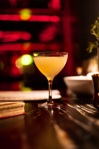 A small crystal glass holding an orange cocktail is on a black bar top. Next to the glass is an old book with the pages worn and tattered. The background is blurred and filled with red fuzzy lighting that gives off a warm and cozy feeling.