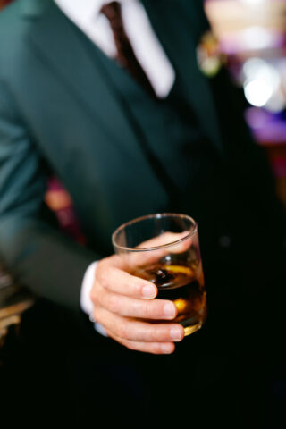 A close-up image of a well-dressed man in a dark green suit, white shirt, and dark tie, holding a glass of whiskey with ice. The background is softly blurred, creating a sophisticated and elegant atmosphere, suggesting a formal event or celebration.