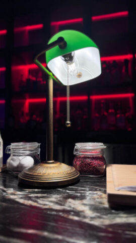 A bankers lamp illuminates a bar top made of black marble swirling with rich brown. In the corner a wooden board is posed surrounded by jars of specialty drink toppings.